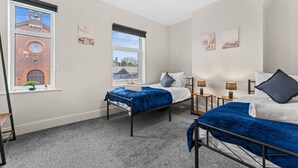 A hotel room with two beds, a window with a view of a brick building, and a small plant on the windowsill. - Stoke Work Stay - Contractors & Families - Sleep 6 (Stoke-on-Trent)