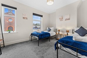 A hotel room with two beds, a window with a view of a brick building, and a small plant on the windowsill. - Stoke Work Stay - Contractors & Families - Sleep 6 (Stoke-on-Trent)