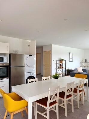 Une cuisine moderne avec un plan de travail blanc, des appareils en acier inoxydable et un coin repas comprenant une table blanche et des chaises jaunes.