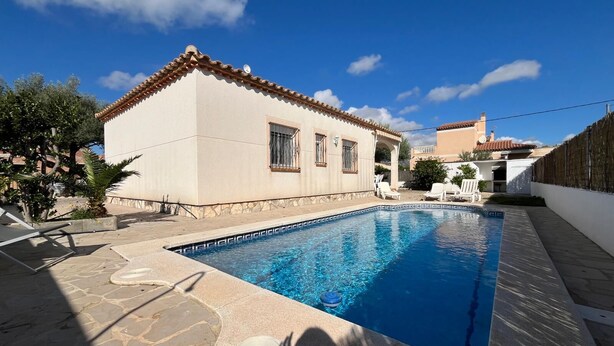 A swimming pool with a stone deck, a white building with a tiled roof, and a clear blue sky. - Villa Esther (Les Tres Cales)