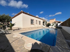 A swimming pool with a stone deck, a white building with a tiled roof, and a clear blue sky.