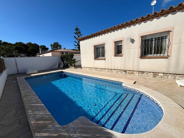 A rectangular swimming pool with blue tiles and a stone border, adjacent to a building with a tiled roof and windows.