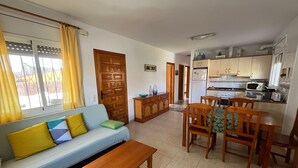 A compact living space with a sofa, a wooden bench, a dining table with chairs, a kitchen area with a refrigerator, microwave, and toaster. - Villa Esther (Les Tres Cales)