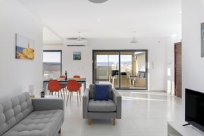 A modern living room with a grey sofa, a grey armchair with a blue pillow, a dining area with red chairs, and a TV on a stand. - Modern Penthouse with Terrace & Sea View (St. Paul's Bay)