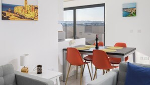 Ein modernes Wohnzimmer mit zwei grauen Sofas, einem Esstisch mit orangefarbenen Stühlen und Blick auf die Stadtlandschaft durch ein großes Fenster.