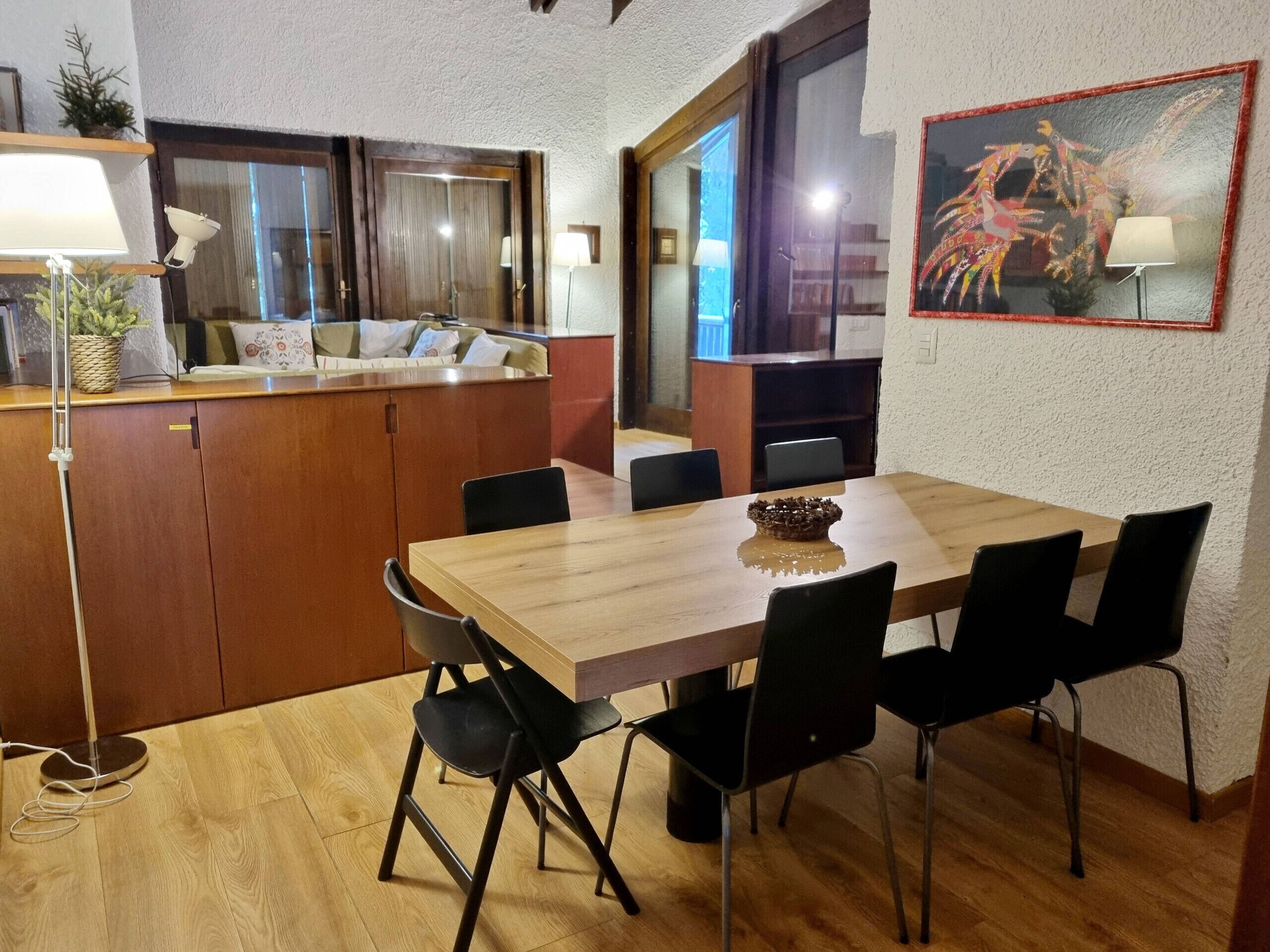 Een eetgedeelte met een houten tafel en zwarte stoelen, een keuken met houten keukenkastjes, en een schilderij aan de muur.