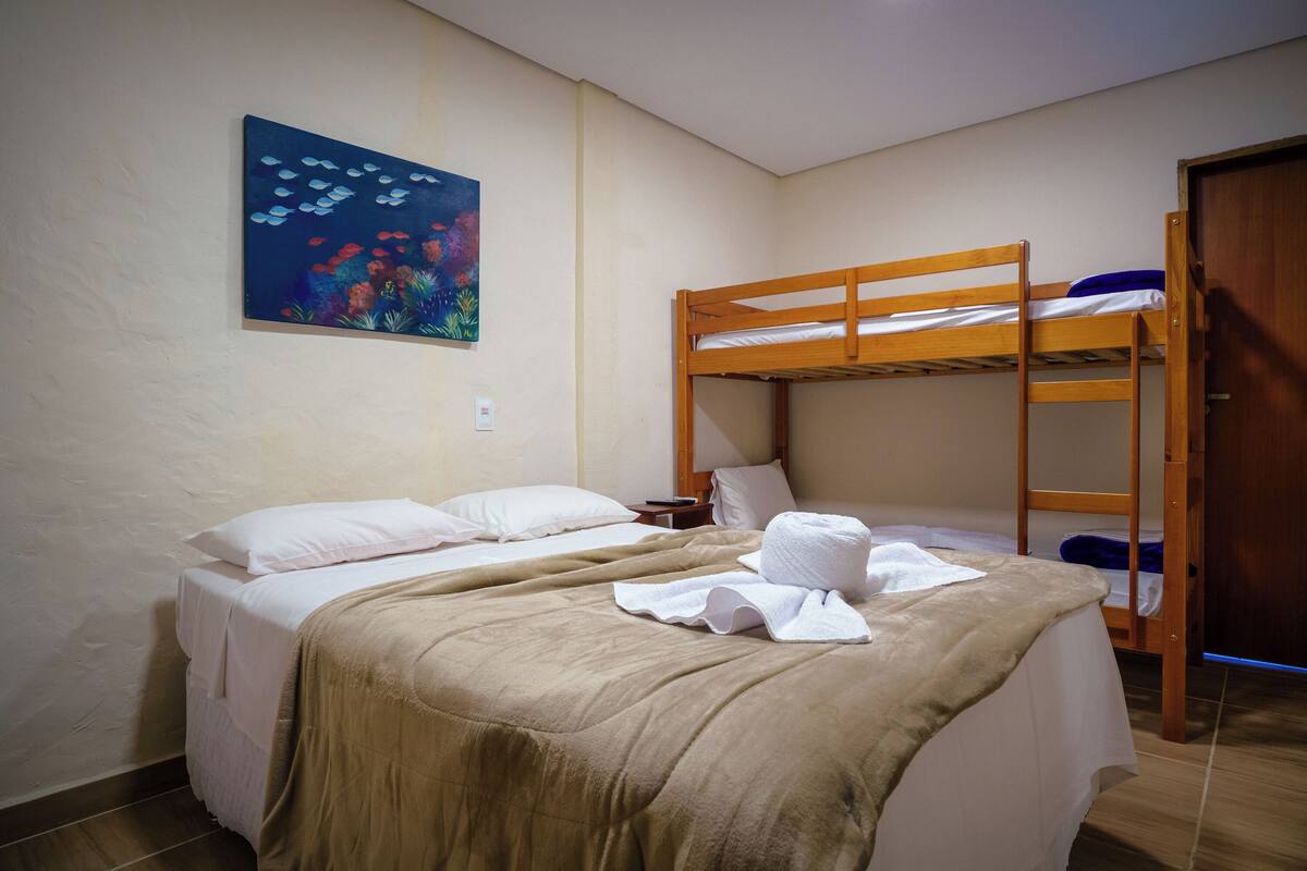 Une chambre avec deux lits, dont un recouvert d’une couverture et de coussins beiges, et un lit superposé avec une couverture bleue. Il y a un tableau représentant des poissons sur le mur et une porte en bois.