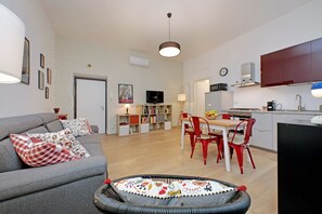 Ein modernes Wohnzimmer mit einem grauen Sofa, roten und weißen Kissen mit Muster, einem Korbstuhl, einem Essbereich mit einem Holztisch und roten Stühlen, einer Küche mit roten Schränken und einem Fernseher auf einem Lowboard.