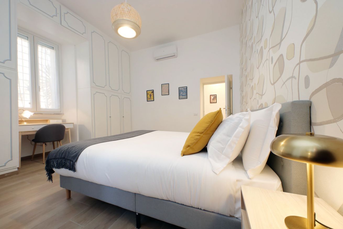 A bedroom with a bed, bedside table, lamp, desk, chair, and window.