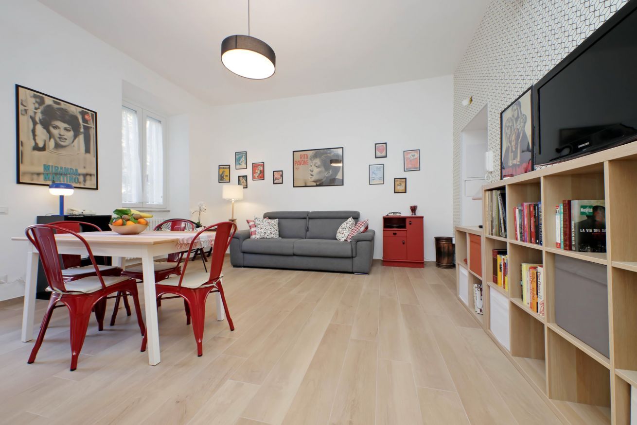 A modern living room with a grey sofa, red chairs, a dining table with a bowl of fruit, and a bookshelf filled with books.