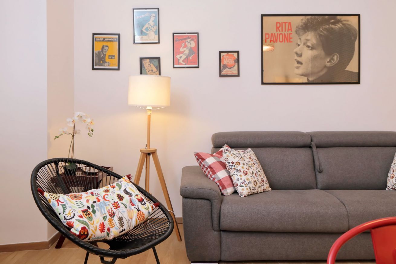Un soggiorno con un divano grigio, una poltrona nera con un cuscino colorato, una lampada da terra e un muro decorato con poster incorniciati.
