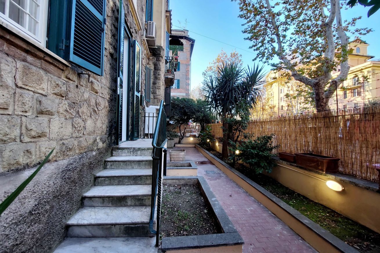 A narrow alleyway with stone steps leading to a building entrance, surrounded by greenery and a bamboo fence.
