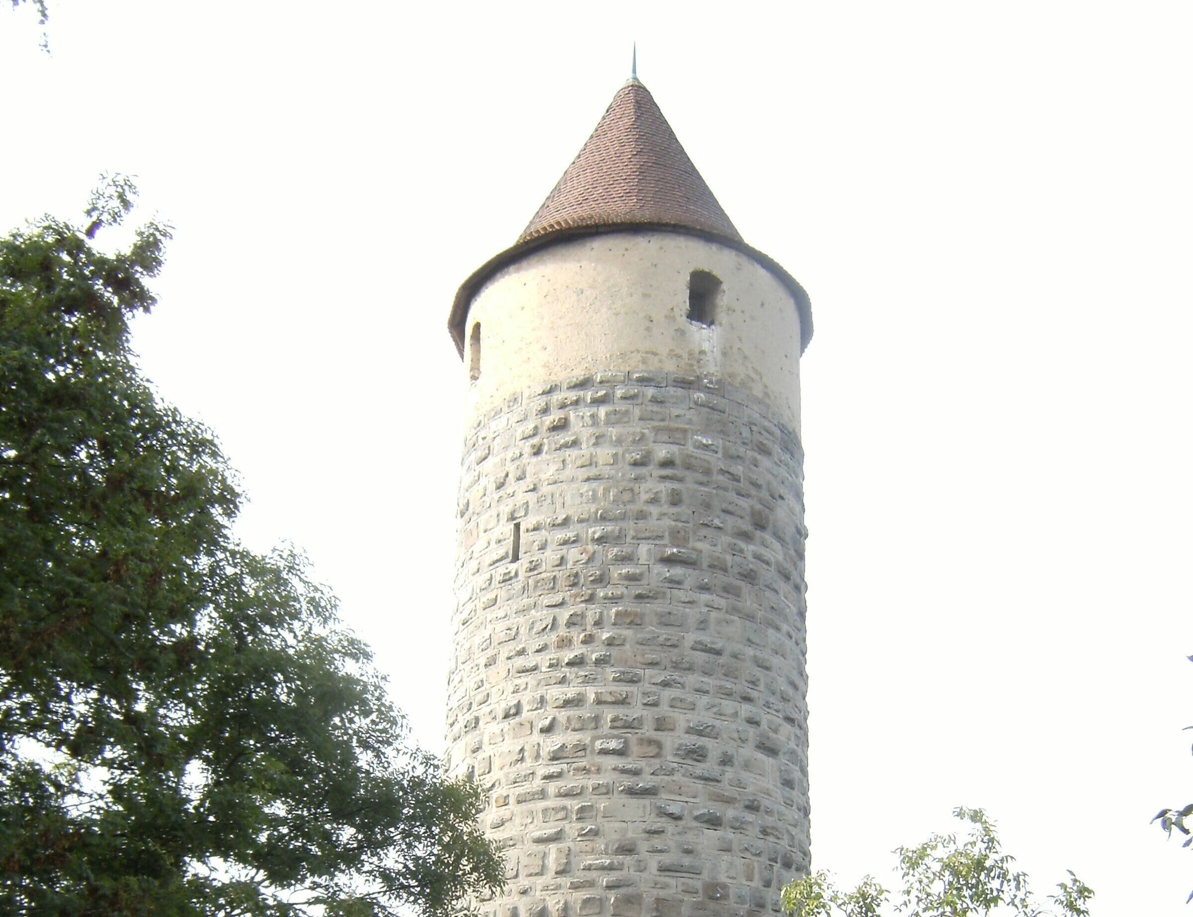 Une haute tour de pierre cylindrique, surmontée d’un toit conique, et entourée d’arbres.