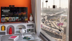 A modern living room with a large window offering a city view, a mounted TV displaying 'Stranger Things', and a bookshelf with books and decorative items. - StayBee - The Artists Gateway Room DownTown Cairo 5mins from Egyptian museum (cairo)