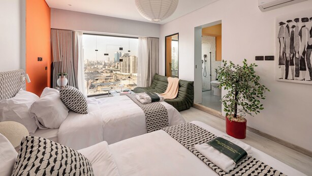 A modern hotel room with two beds, a city view, a large window, a plant, and a bathroom visible through an open door. - StayBee - The Artists Gateway Room DownTown Cairo 5mins from Egyptian museum (cairo)