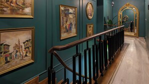 A hallway with teal walls, wooden flooring, and a black railing. There are framed artworks on the walls and a large mirror with a gold frame at the end of the hallway. - StayBee - The Artists Gateway Room DownTown Cairo 5mins from Egyptian museum (cairo)