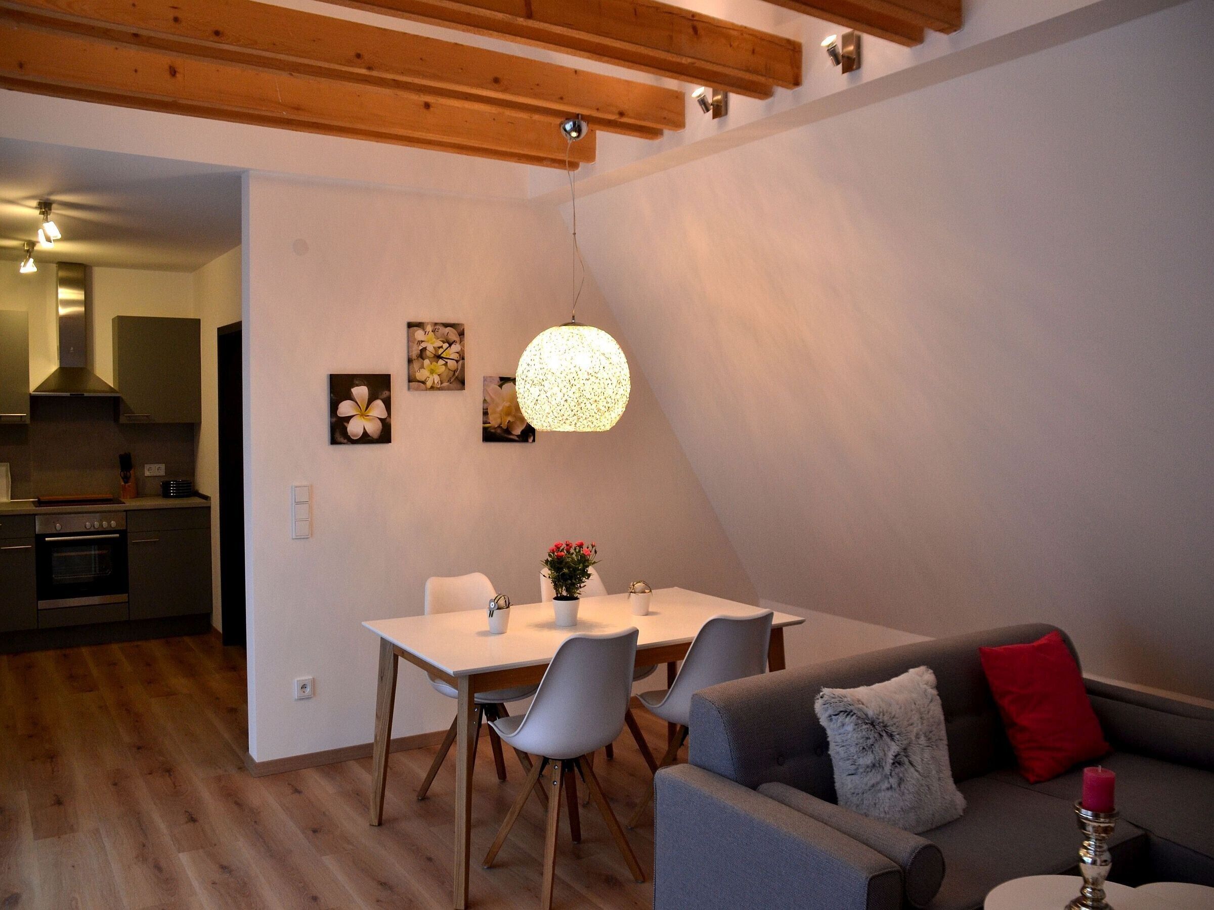 A modern kitchen with wooden beams, a dining area with a table and chairs, and a living room with a sofa and a red pillow.