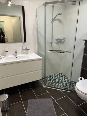 Uma casa de banho moderna com chuveiro em cabine de vidro, lavatório branco com espelho e soalho em mosaico escuro.