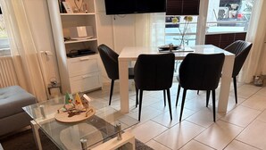 Un salon moderne avec une table basse en verre, un canapé, une table à manger avec des chaises, un téléviseur et une vue sur l’extérieur par une fenêtre.
