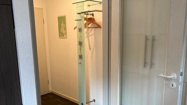 Un couloir avec une porte donnant sur une pièce, une cabine de douche en verre et un porte-manteau avec des cintres.