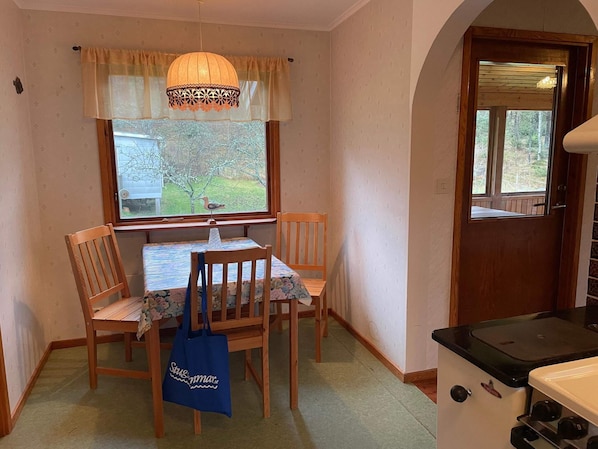 En matplats med ett bord och stolar, ett fönster med utsikt över träd, en hängande lampa och ett köksområde med en spis.