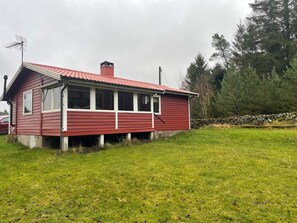 Uma casa de madeira vermelha com telhado inclinado, chaminé e um muro de pedra ao fundo.