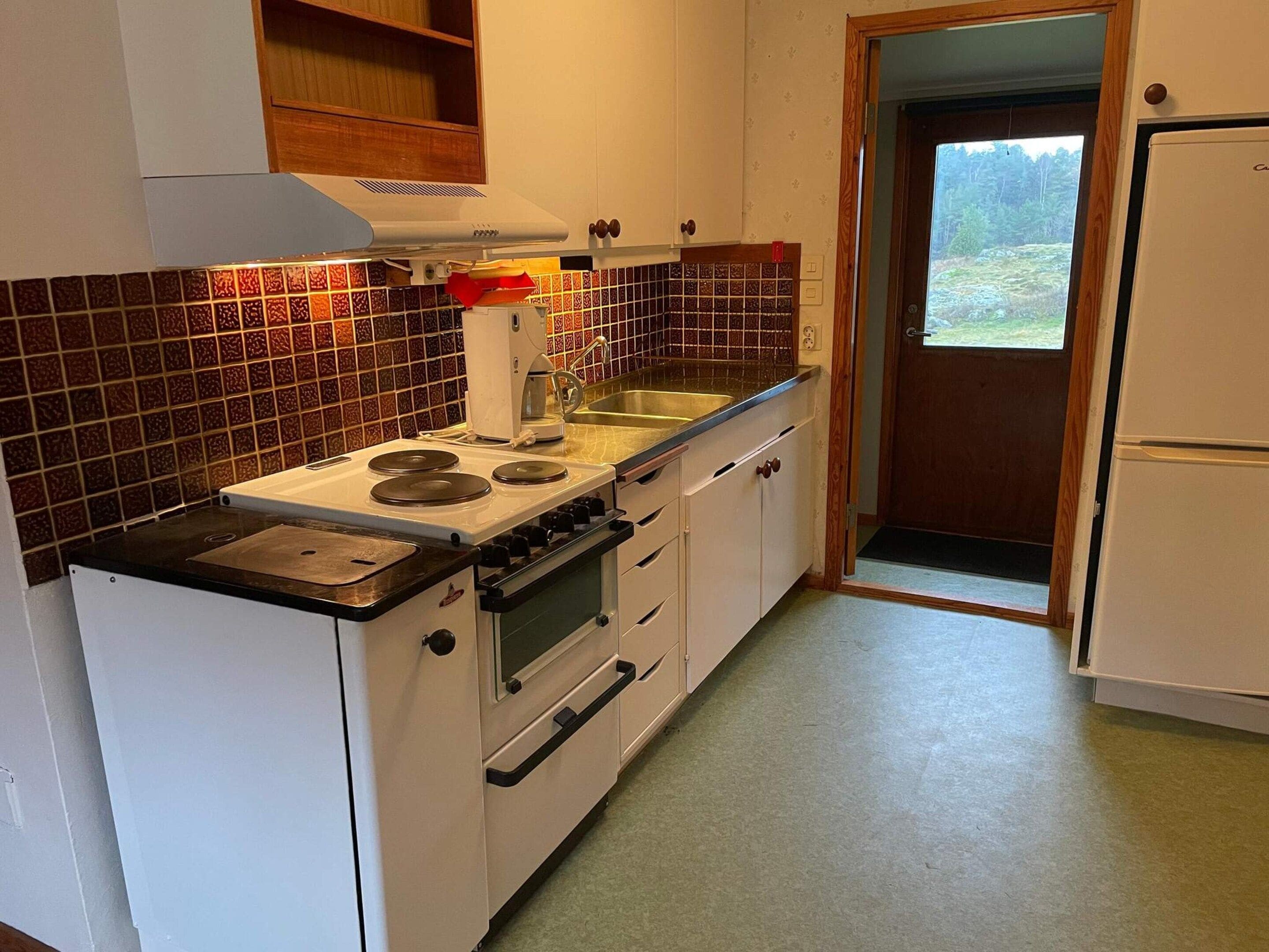 Cozinha compacta com fogão, forno e pia. Há um armário de madeira acima do fogão e uma porta que dá para uma varanda.