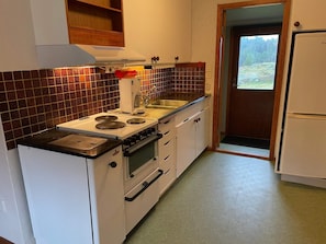 Ett kompakt kök med en spis, ugn och diskho. Ovanför spisen finns ett träskåp och en dörr som leder ut till en balkong.