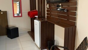 A compact room with a television mounted on a wooden wall, a small refrigerator, a wicker chair, and a red-framed mirror. - Pousada Rota do Parque (Penha)
