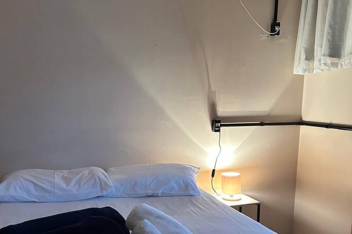 A neatly made bed with white linens and a dark blanket, a bedside lamp, and a wall-mounted air conditioning unit.