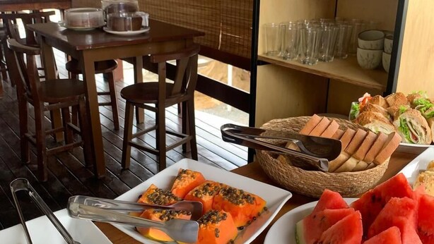 A dining table with plates of food, including watermelon slices and a dish with a yellow topping. There are also bread and a basket of utensils on the table. - Pousada Rota do Parque (Penha)