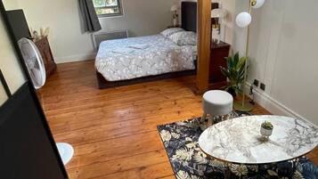 Een kleine slaapkamer met dakraam, een bed, nachtkastje, klein rond tafeltje met een plant en een ventilator.