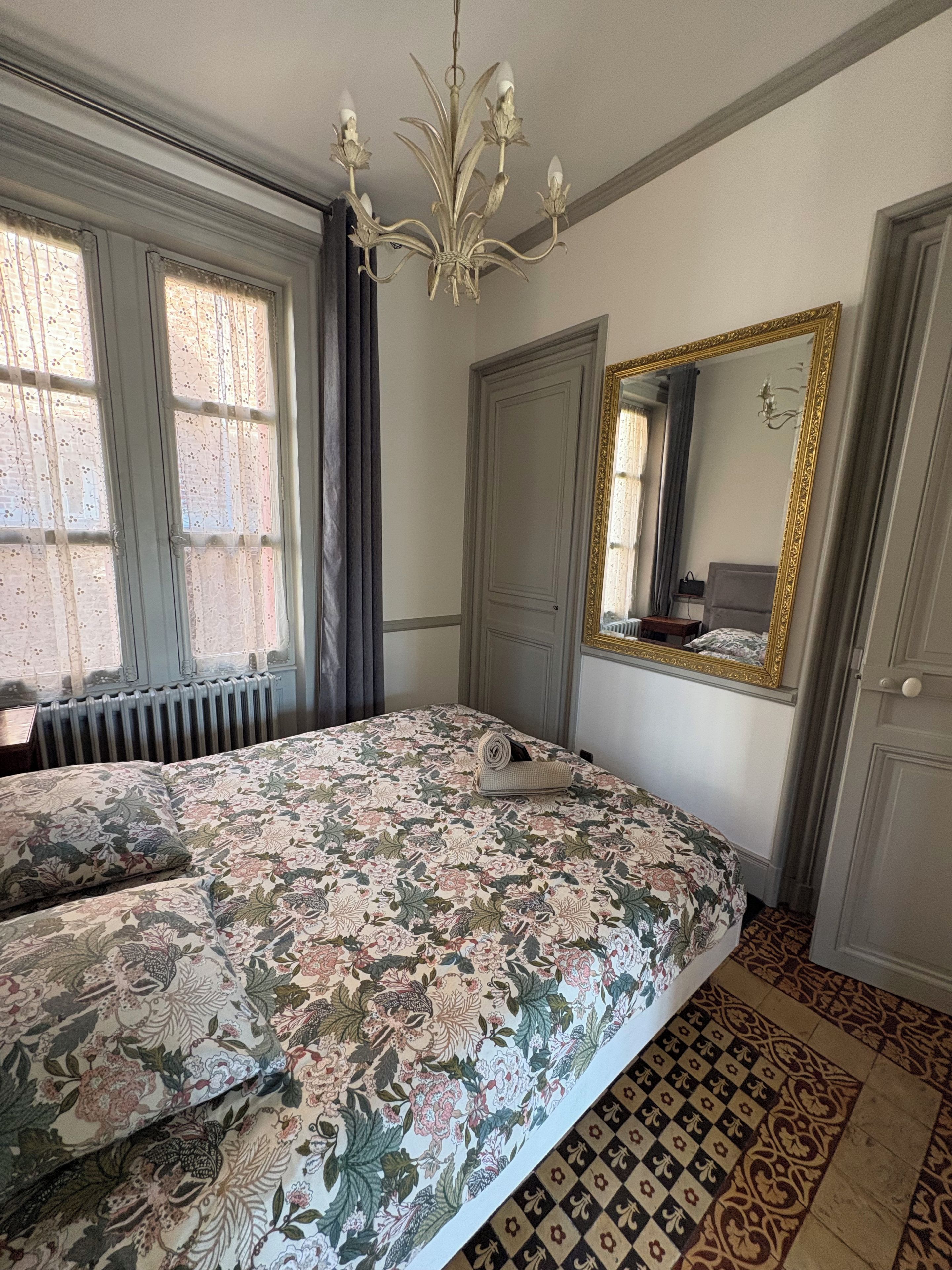 Un dormitorio con colcha estampada, un candelabro, un espejo grande con marco dorado y suelo con diseño.