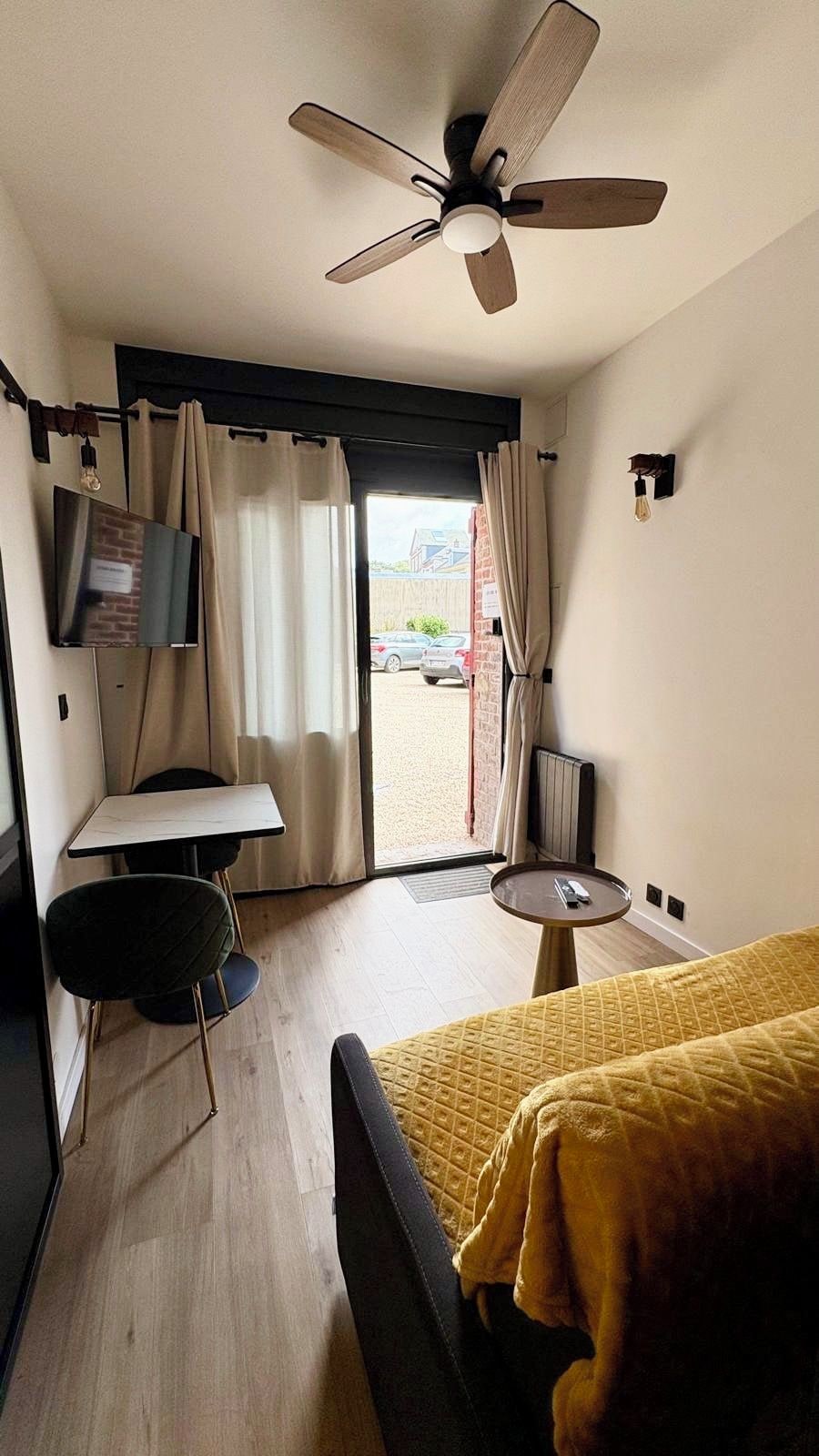 A hotel room with a bed, a small table, a chair, a ceiling fan, a TV, and a view of the outside.