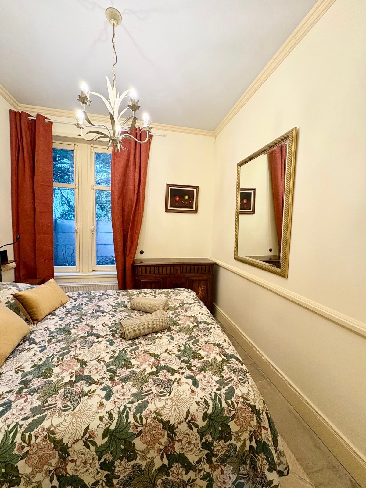 Un dormitorio con cubrecama floral, una araña, un espejo grande y una ventana con cortinas.