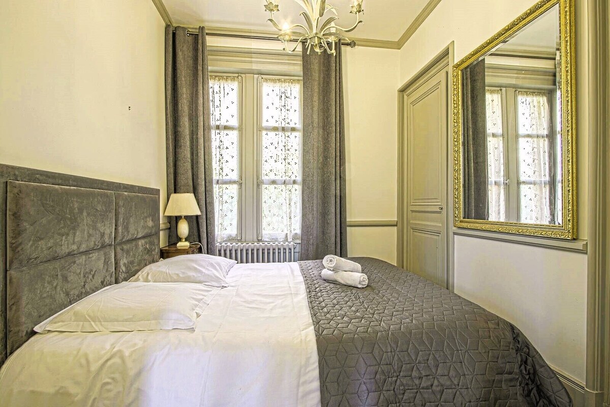 A neatly made bed with a headboard, a nightstand with a lamp, a window with curtains, and a large mirror with a decorative frame.
