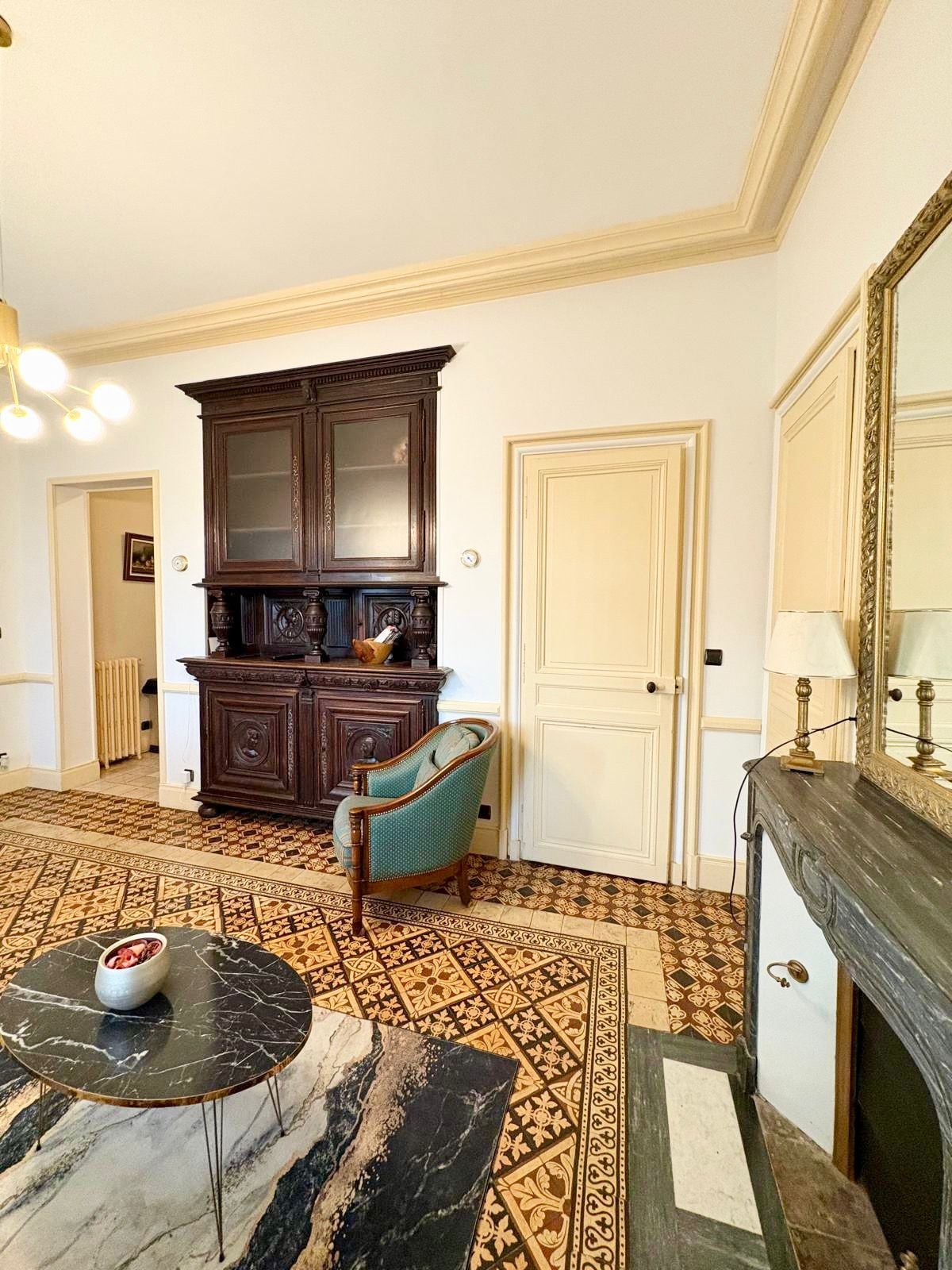 A room with a patterned carpet, a dark wooden cabinet, a blue armchair, a marble table, and a white door.