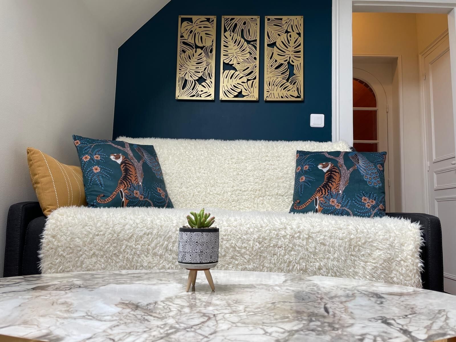 A living room with a dark blue accent wall, a white fluffy sofa, two tiger-themed pillows, a small potted plant, and a marble coffee table.