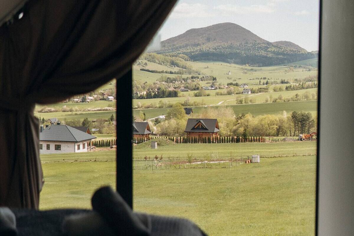 Un dormitorio con una ventana grande, una cama y vistas a un paisaje rural con casas y una colina.