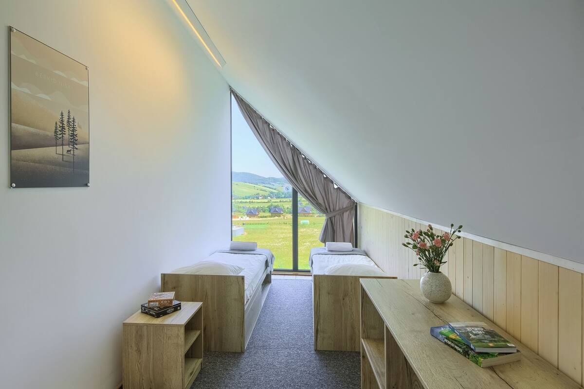 Una habitación con dos camas, un banco de madera, una mesita, un jarrón con flores y un ventanal con vistas a un paisaje verde.