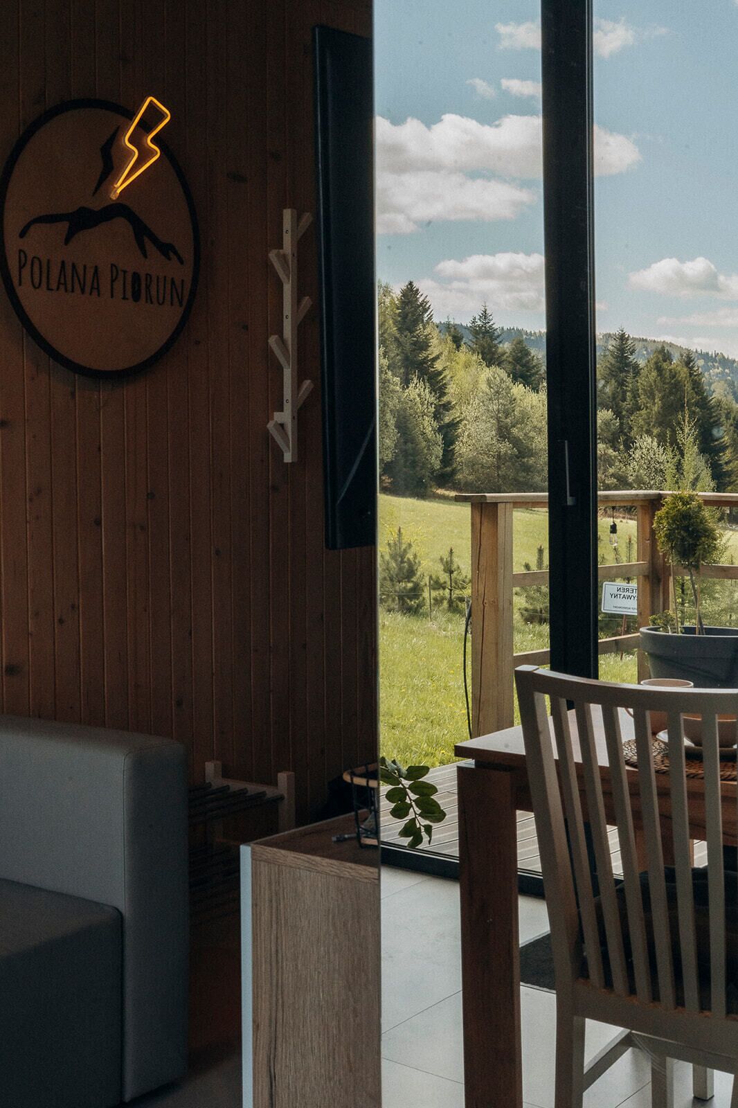 A room with a wooden wall, a sign reading 'POLANA PIORUN,' a view of a green landscape through large windows, and a wooden table with a chair.