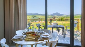 Un tavolo da pranzo con piatti, bicchieri e cibo, affacciato su una zona esterna panoramica con vista su colline e una casa.