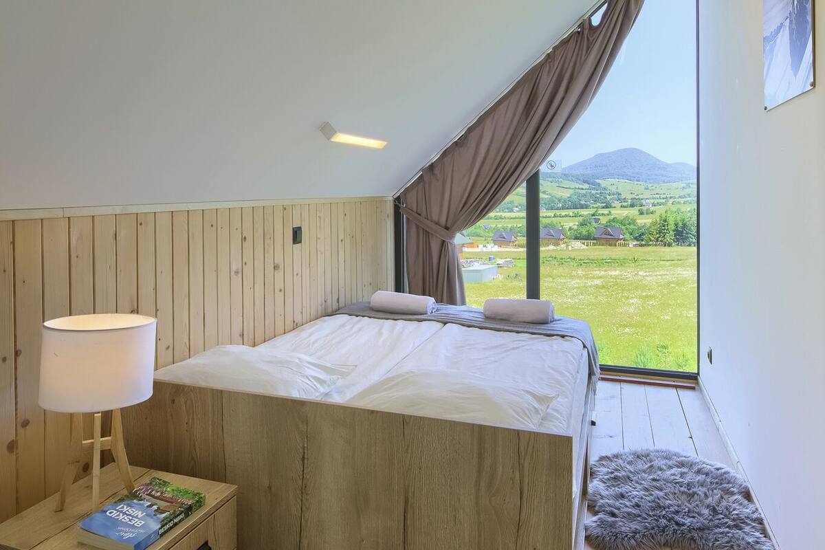 Un dormitorio con cabecera de madera, una mesita de noche con lámpara, una alfombra y un gran ventanal con vistas al campo.