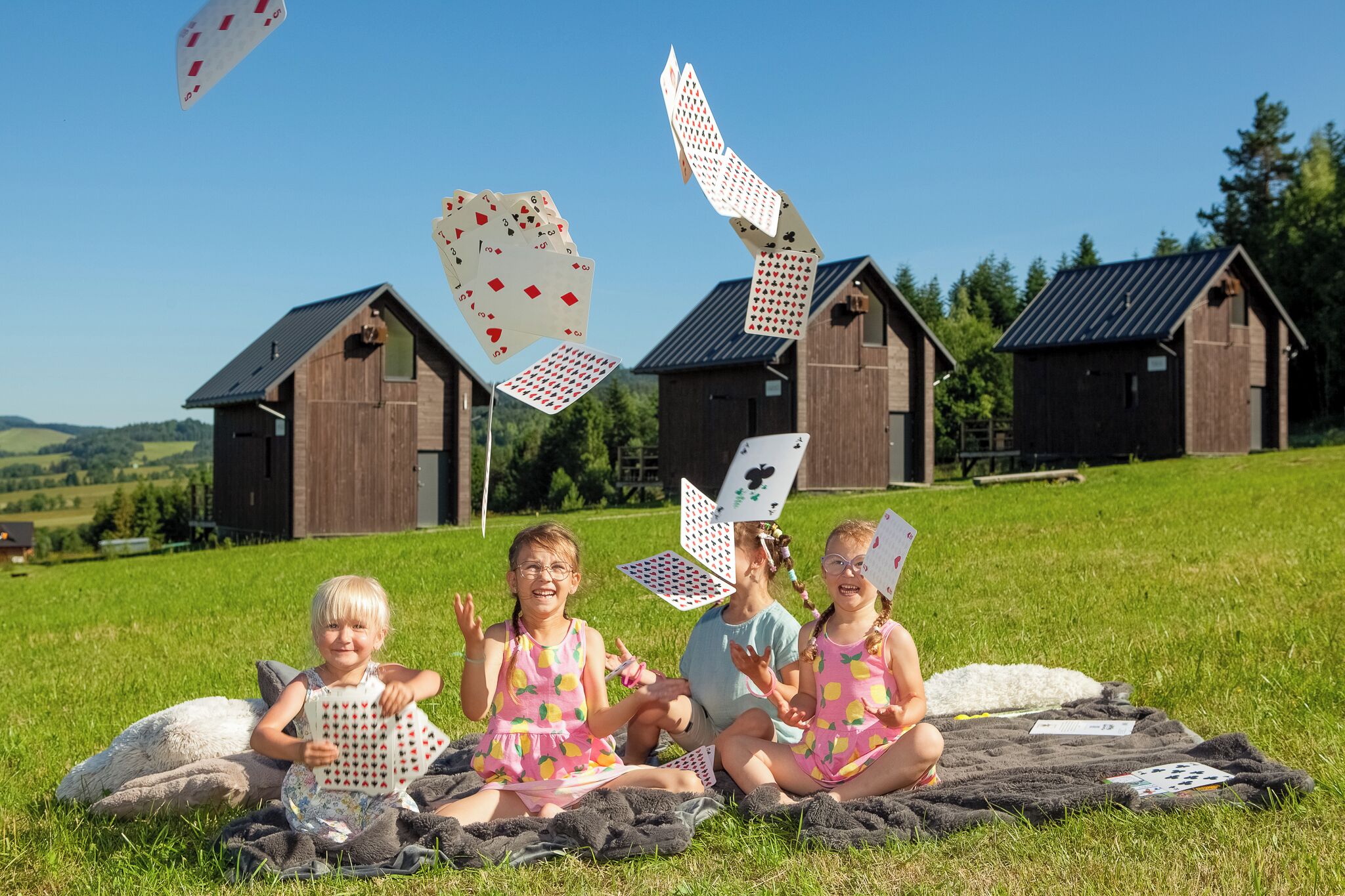 Des enfants jouent aux cartes sur une couverture, dans un champ herbeux, avec des granges en arrière-plan.