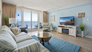 A living room with a sofa, armchair, and a glass coffee table. There is a television on a stand, a plant, and a view of the ocean through the balcony doors. - Two-bedroom oceanfront with indoor and outdoor pools! (Ocean City)