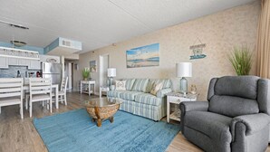 A living room with a sofa, armchair, and coffee table. There is a kitchen area with a refrigerator and cabinets. - Two-bedroom oceanfront with indoor and outdoor pools! (Ocean City)