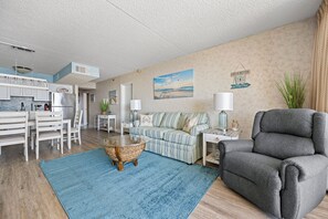 Ein Wohnzimmer mit einer Couch, einem Sessel und einem Couchtisch. Es gibt eine Küchenzeile mit einem Kühlschrank und Schränken.