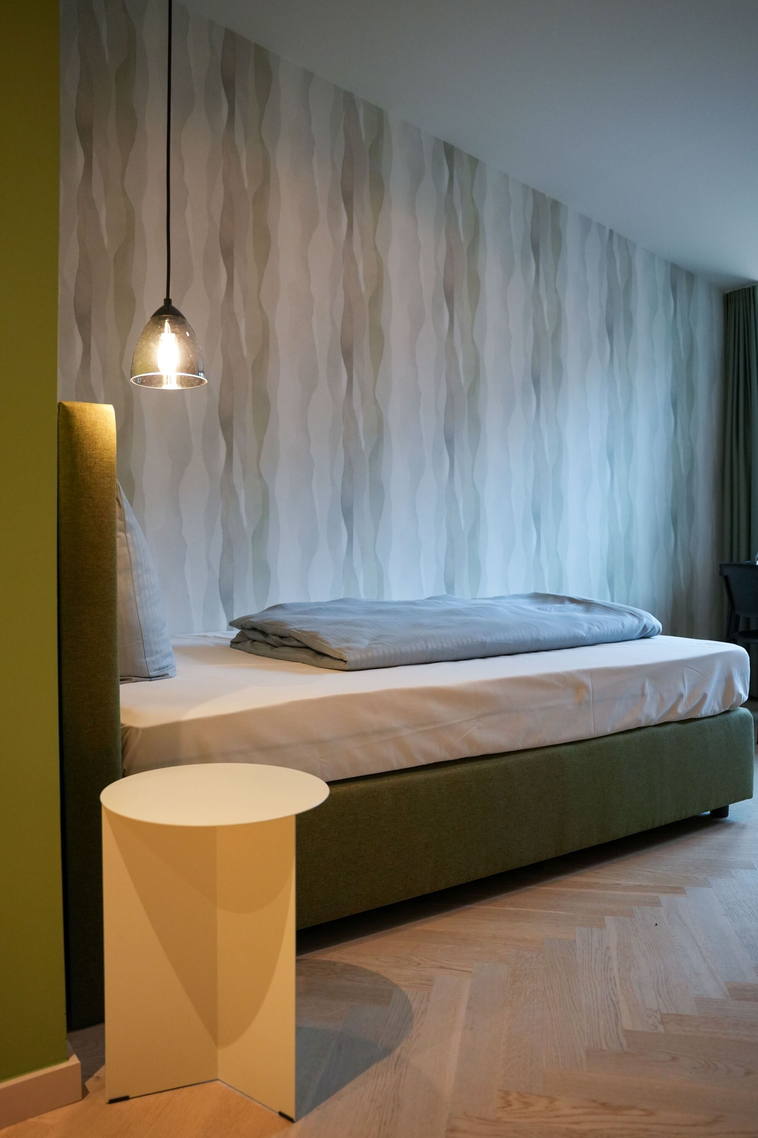 A modern bedroom with a bed, a round side table, a textured wall, and a hanging light fixture.
