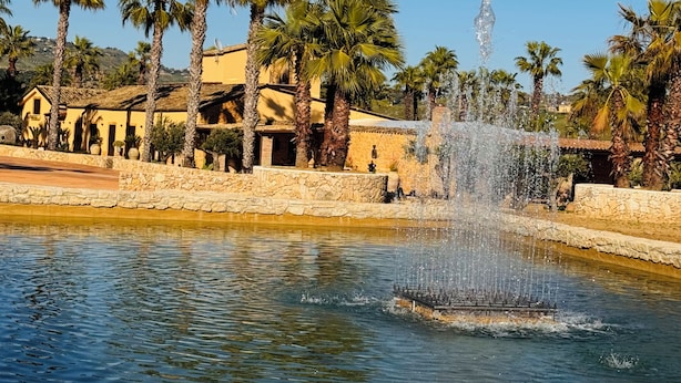 A fountain in a courtyard with palm trees and a building with yellow walls. - Villa Demetra Resort (Agrigento)