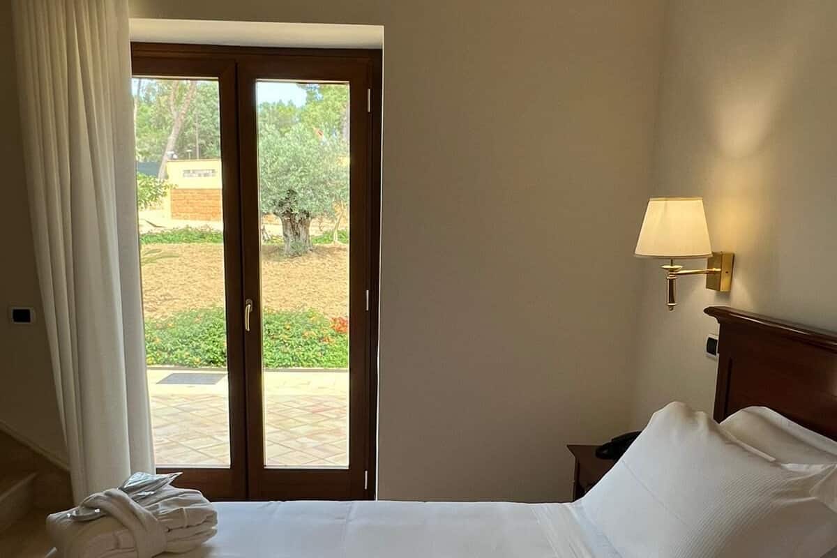 Une chambre d’hôtel avec un lit, une table de chevet, une lampe, une fenêtre avec des rideaux et une vue sur la verdure.